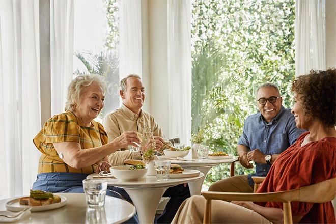 Almoço em família, dois casais sentados com uma mesa e comida entre eles. A senhora a esquerda da foto, vestindo uma camisa em tons amarelos possui o Sensor FreeStyle Libre no braço direito.