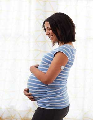 Mulher grávida sorrindo, acariciando a sua barriga, com um sensor de glicemia no seu braço esquerdo.