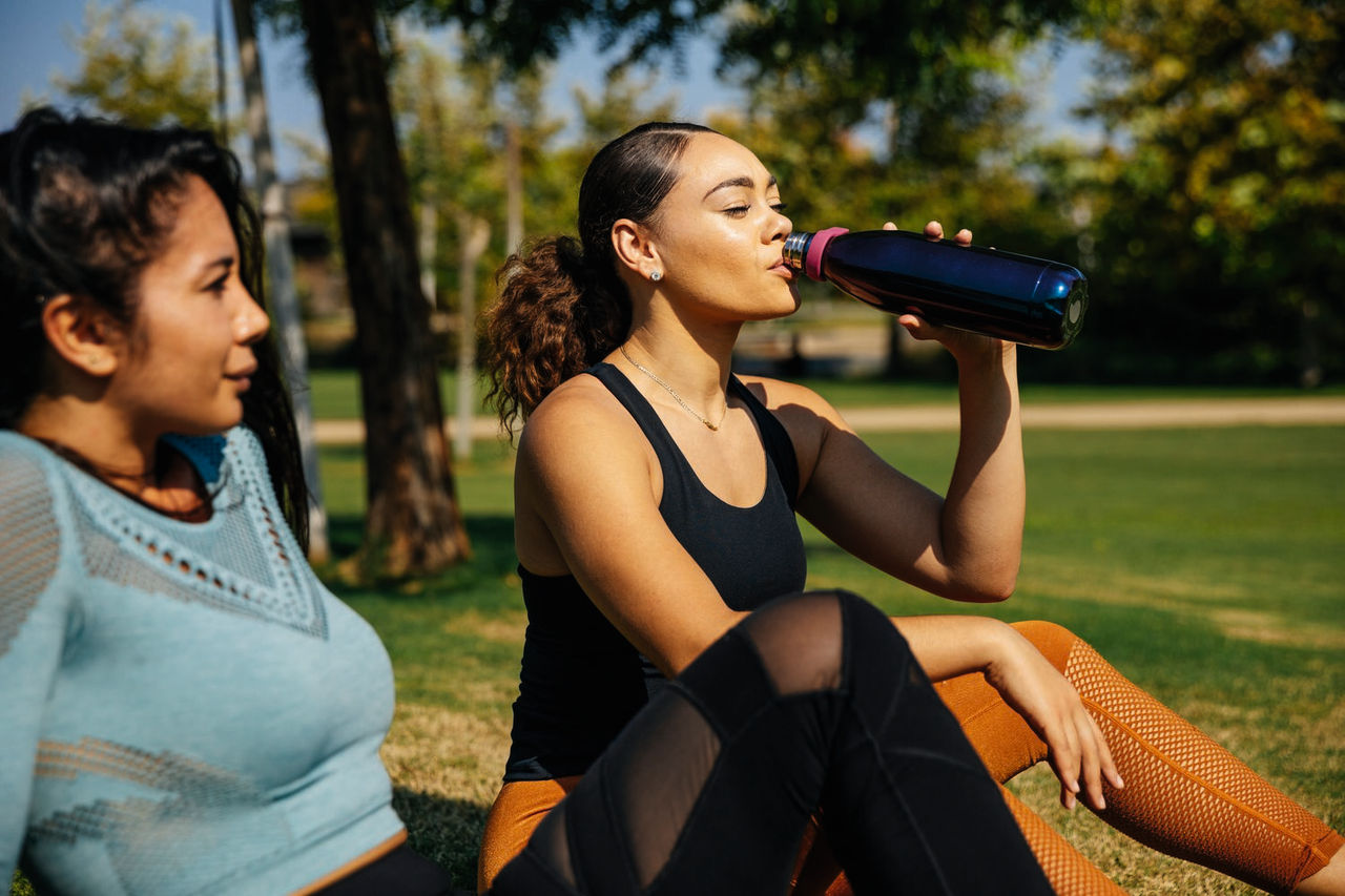 Young woman drinking water while sitting with female friend at park - stock photo
